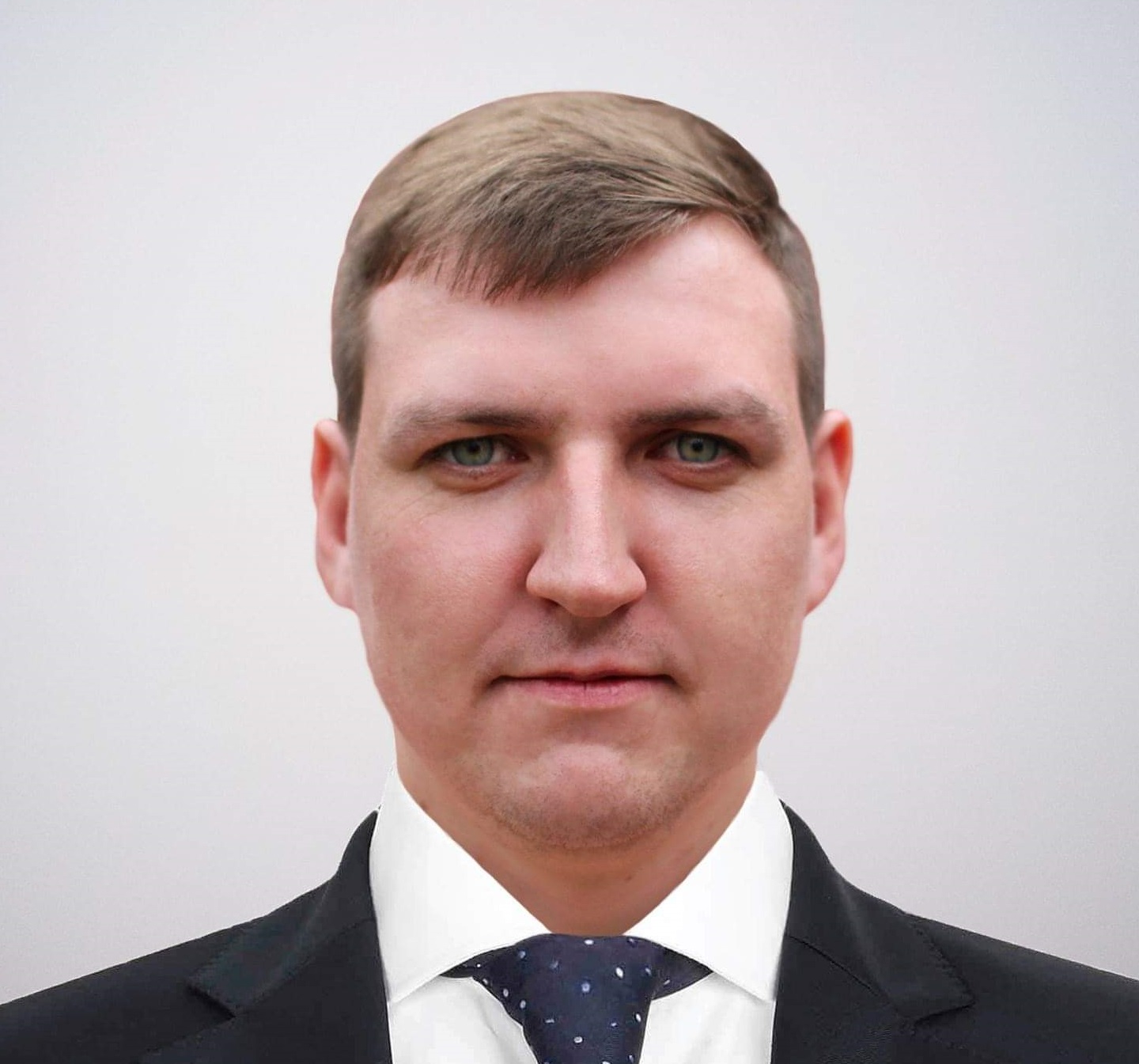 Portrait of a man with short brown hair, wearing a black suit, white shirt, and a dotted blue tie, set against a pale background.