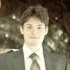Portrait of a smiling young man in a suit with a tie, standing outdoors with a blurred natural background.