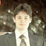 Portrait of a smiling young man in a suit with a tie, standing outdoors with a blurred natural background.