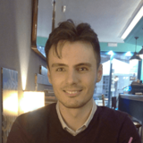 A smiling young man with styled hair, wearing a dark sweater over a collared shirt, in an indoor setting with soft lighting and blurred background.