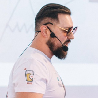 Profile of a man with a beard wearing sunglasses and a headset microphone, speaking at an event, with a blurred background.