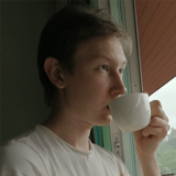 A person in a white t-shirt drinking from a white cup, looking out a window, with a contemplative expression.