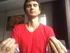 Man in red v-neck shirt meditating with eyes closed, hands in a mudra position, in a bedroom with a flower painting in the background.