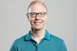 Smiling person with glasses wearing a teal polo shirt against a gray background.