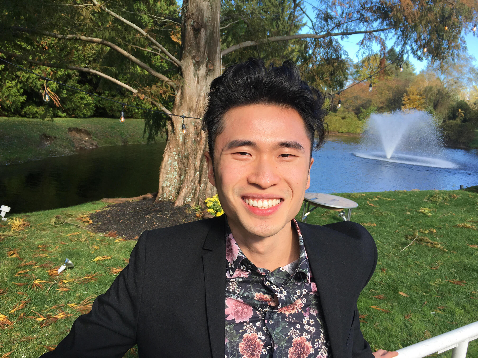 A smiling man in a suit with a floral shirt standing outdoors with a fountain in a pond and autumn foliage in the background.