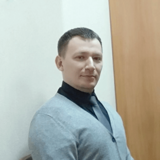 Man in a grey blazer and black turtleneck smiling at the camera in an indoor setting.