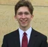 Young man with a cheerful smile wearing a black suit and red tie standing before a beige wall.