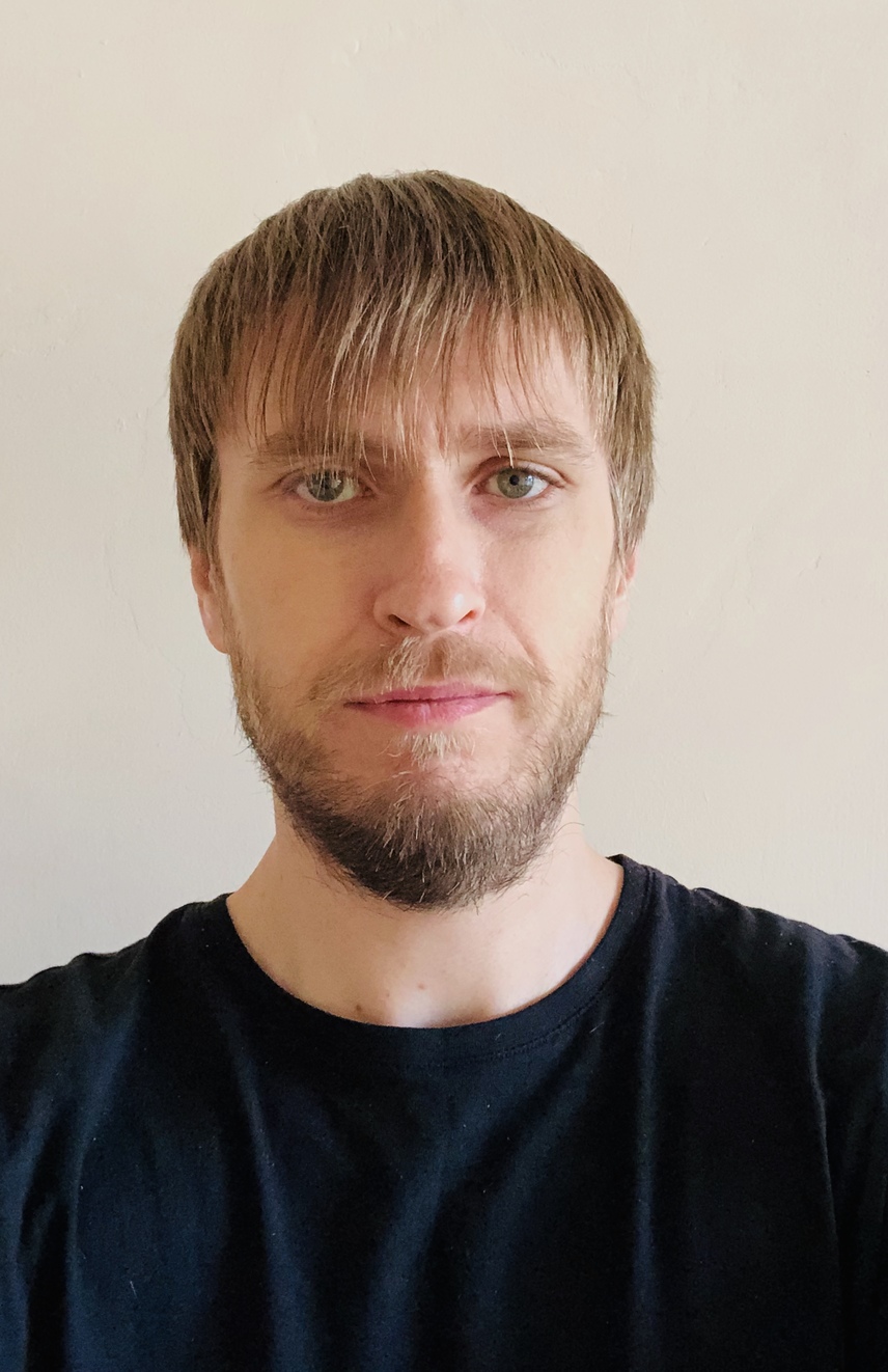 A headshot of a man with short brown hair, a beard, and blue eyes, wearing a black t-shirt against a light-colored wall background.