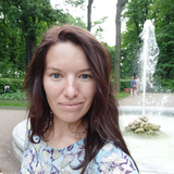 A woman with shoulder-length brown hair smiling at the camera with a fountain in the background and trees surrounding the area, indicating a park setting.