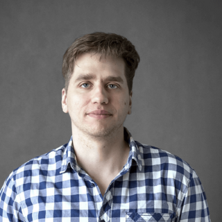 Portrait of a smiling young man with light brown hair wearing a blue and white checked shirt. The background is a neutral gray color.