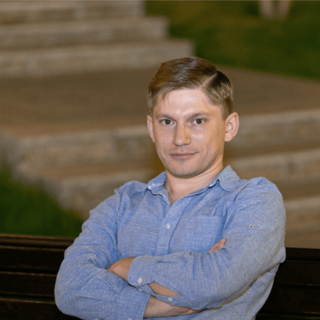 A man in a blue shirt sitting on a bench with arms crossed, smiling slightly at the camera, with soft-focus stairs in the background.