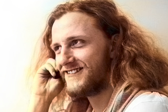 A smiling man with long wavy hair and a beard talking on a mobile phone, with a pleasantly blurred background providing a clear focus on the subject.