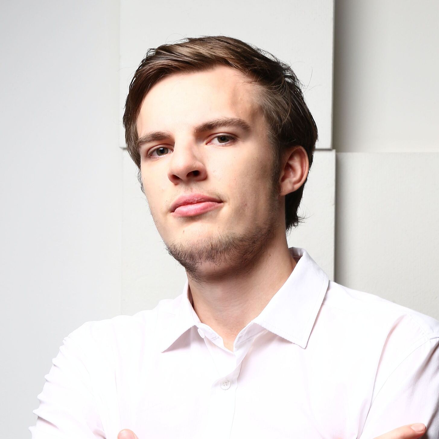 Headshot of a young man with a slight beard wearing a white shirt, standing against a white backdrop, looking at the camera with a serious expression.