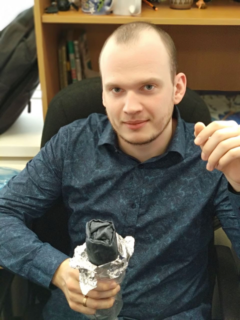 A man with a shaved head, wearing a blue patterned shirt, sitting at a desk holding a rose wrapped in foil. Behind him, a shelf with books and figurines.