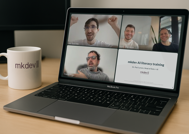 Laptop on desk showing a video conference with three participants and a slide presentation titled "mkdev AI literacy training." A white mug with "mkdev" branding is beside the laptop.