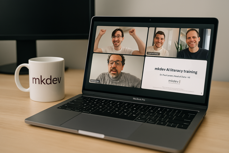 Laptop screen showing a virtual meeting with three participants and a slide titled "mkdev AI literacy training." A white mug with "mkdev" on it sits next to the laptop on a wooden desk.